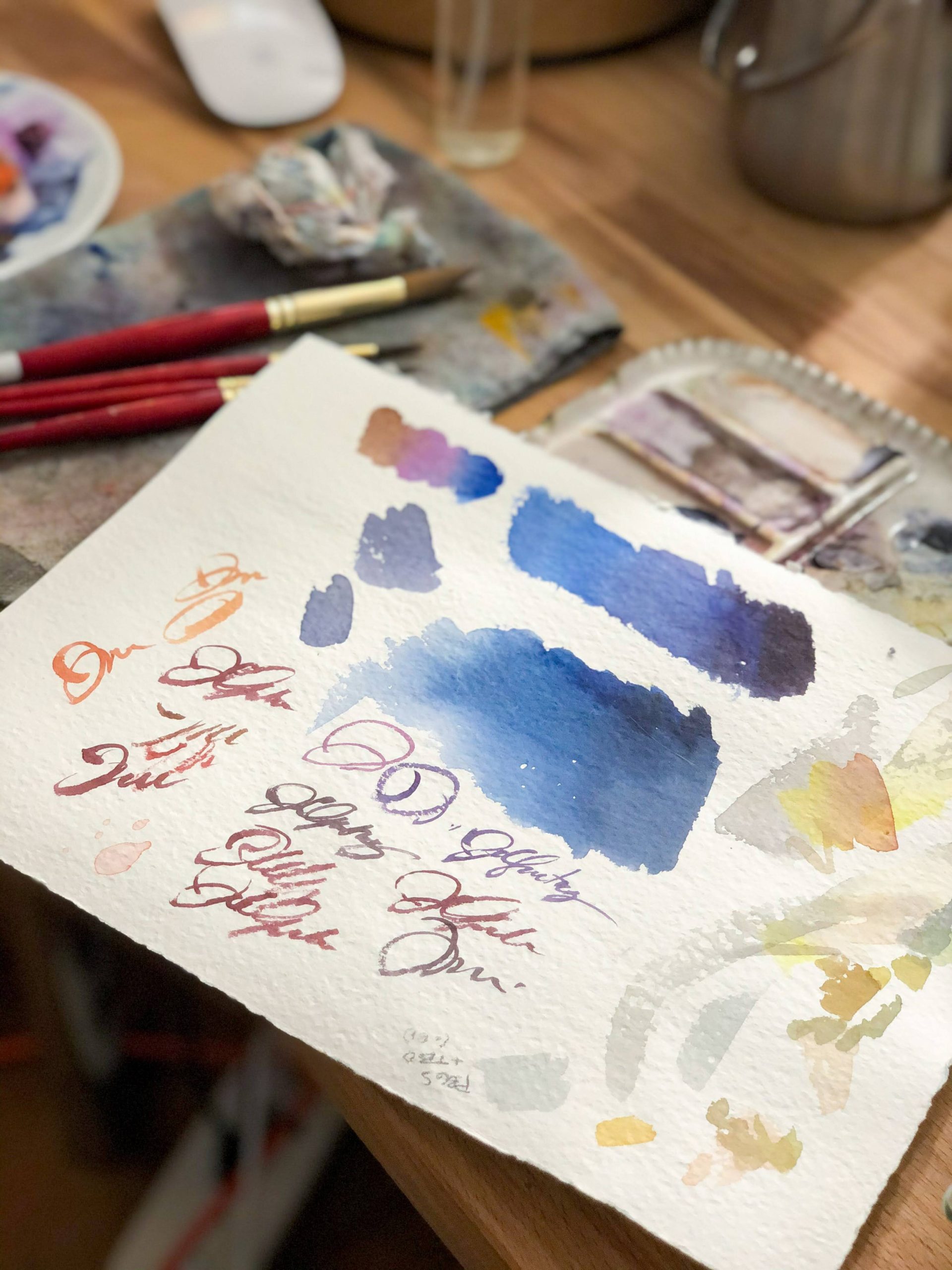 Foreground with paper covered in swatches and practice signatures. Background is art supplies on desk.