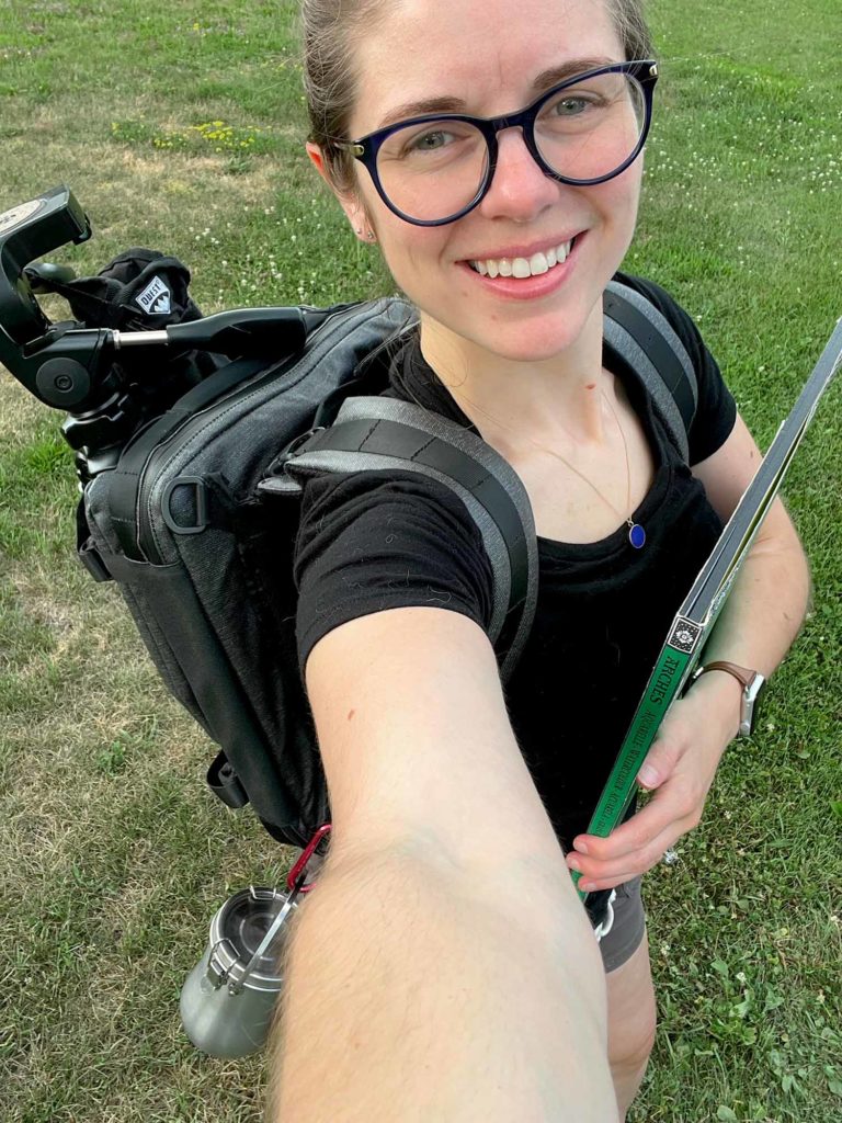 Large Watercolor Plein Air Setup Step  10: Jill has satchel with tripod and water jar on in backpack mode and carries a large block of paper in the hand not holding the camera.