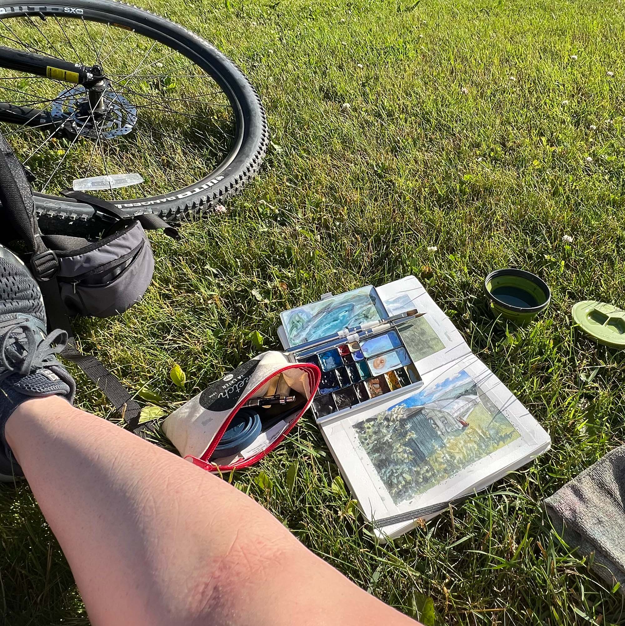 Jill's portable landscape palette and kit on the grass next to her leg and bike wheel. There is a plein air sketch of a local farm in the sketchbook.